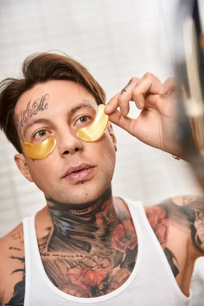 Giovane uomo con tatuaggi gode di un momento accogliente a casa, applicando una maschera calmante occhio d'oro. — Foto stock