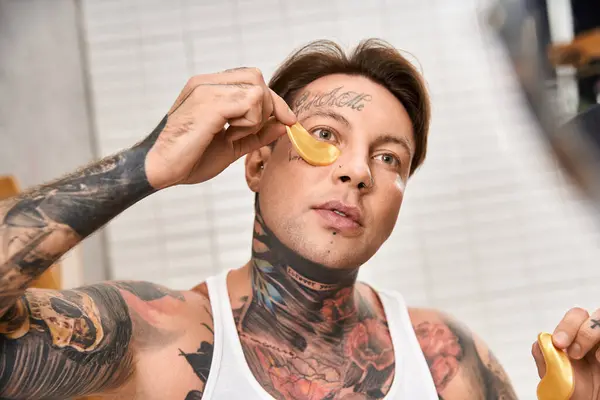 Ein junger tätowierter Mann entspannt sich zu Hause, während er eine beruhigende Behandlung unter seinen Augen anwendet. — Stockfoto