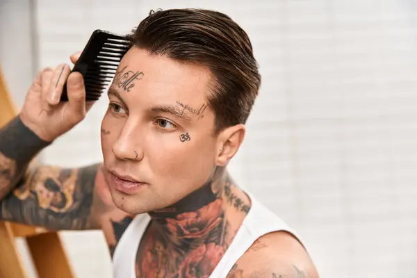 Un hombre joven con tatuajes estiliza su cabello mientras se siente cómodo en casa en un ambiente acogedor. - foto de stock
