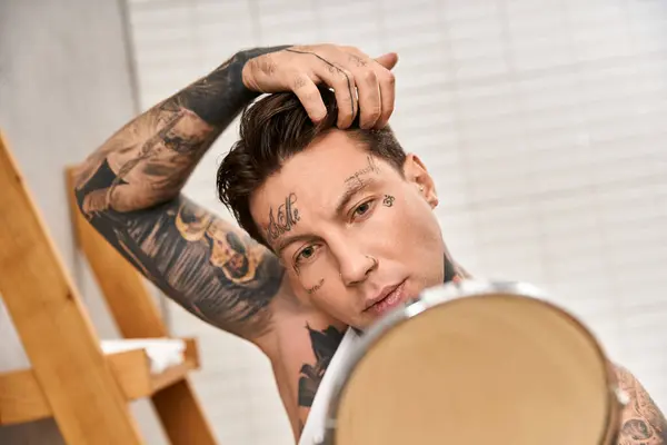 A handsome tattooed man adjusts his hair while looking in a mirror, surrounded by a cozy atmosphere. — Stock Photo