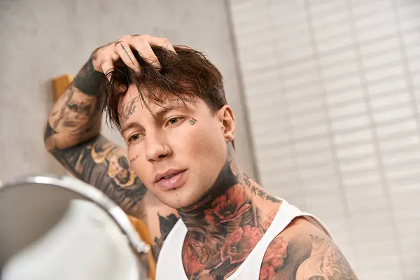 Young tattooed man in a comfy atmosphere looks at his reflection while adjusting his hair. — Stock Photo