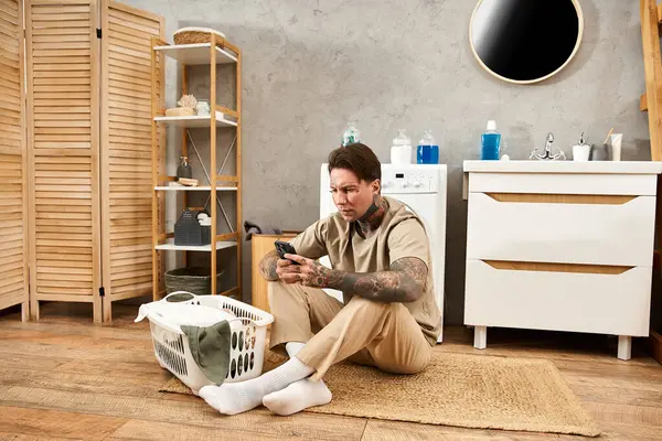 Junger Mann mit Tätowierungen sitzt gemütlich zu Hause und tippt mit seinem Handy in einem gemütlichen Raum. — Stockfoto
