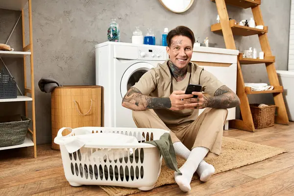Ein tätowierter junger Mann lächelt auf dem Boden, entspannt mit seinem Smartphone in einem gemütlichen Raum. — Stockfoto