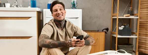 Ein junger Mann mit Tätowierungen lächelt strahlend, während er sich zu Hause entspannt, in sein Handy vertieft. — Stockfoto