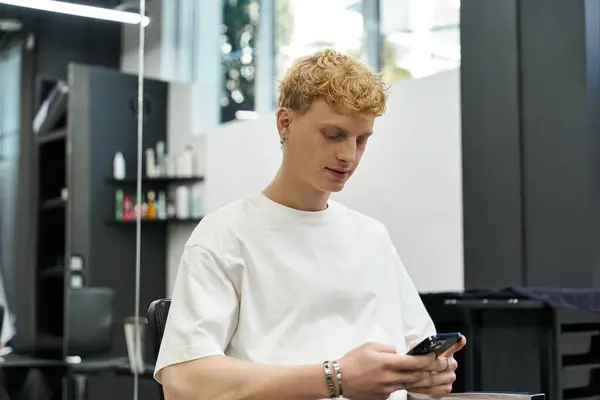 Young man sits in beauty salon chair, focused on his phone, waiting for a haircut. — Stock Photo