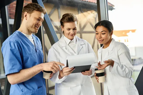 Three multicultural doctors discuss patient care while reviewing notes in a contemporary hospital. — Stockfoto