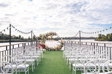 The wedding arch stands in front of a row of chairs on a wooden pier overlooking the bay of the river