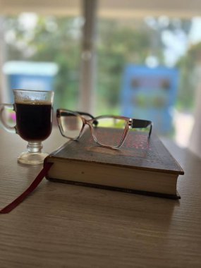 Üstünde bardaklar olan bir kitabın yanında bir fincan sade kahve. Fotoğrafın bulanık arkaplanı