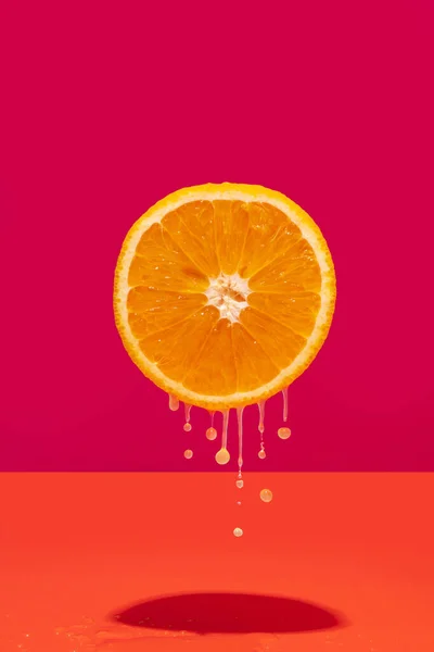 Conceptual levitating half orange with dripping juice on vibrant dark pink and orange background