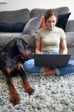 Genç bir kadın evdeki kanepenin önünde oturmuş yanında evcil bir köpekle dizüstü bilgisayarda çalışıyor.                               
