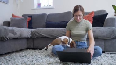 4K videosunda, evinde koltuğun önünde oturan genç bir kadın yanında evcil bir köpekle dizüstü bilgisayarda çalışıyor.