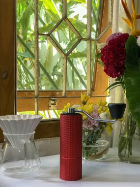 Kırmızı bir el yapımı kahve öğütücüsü arka planda çiçekler ve yemyeşil bir pencerenin yanındaki masada oturur..