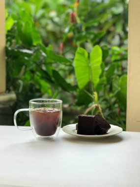 Bir bardak buzlu kahve ve bir dilim çikolatalı kek, yeşillik manzaralı beyaz bir masanın üzerinde..