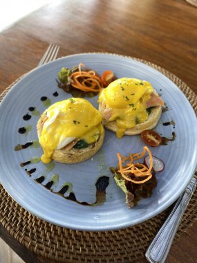 İngiliz kekleri üzerine füme somon, ıspanak ve kremalı hollandaise soslu iki yumurtalı Benedict, mavi bir tabakta salata ile servis ediliyor.