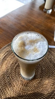 Bir bardak buzlu kahve ile latte sanatı, ahşap bir masanın üzerinde dinleniyor, yanında iki sürahi süt veya krema var..