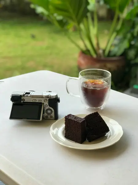 Açık havada beyaz bir masa, bir bardak buzlu kahve, bir tabak çikolatalı kek ve ekranı açık olan aynasız bir kamera..