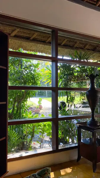 Ahşap çerçeveli büyük bir pencere, patikası, bitkileri ve geleneksel çatısı olan yemyeşil bir tropikal bahçenin huzurlu bir görüntüsünü sunuyor..