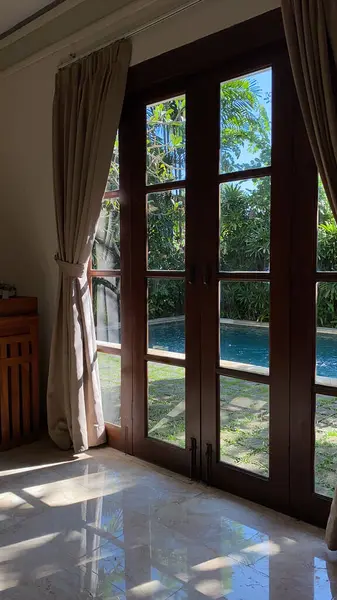Açık Fransız kapılarını çerçeveleyen bej perdeli güneşli bir oda, yemyeşil bir bahçe ve yüzme havuzu manzarası ortaya çıkarıyor..