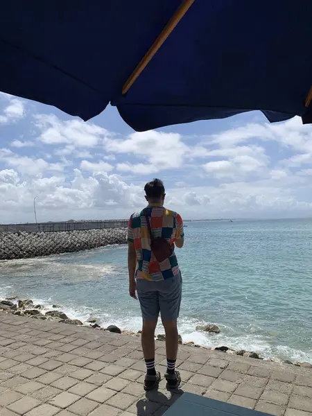 Sahilde şemsiyenin altında duran bir adam var. Sahilde şemsiye altında duran adam; seyahat broşürleri, hava durumu içeriği, tatil reklamları, açık hava eğlence promosyonları için uygun.