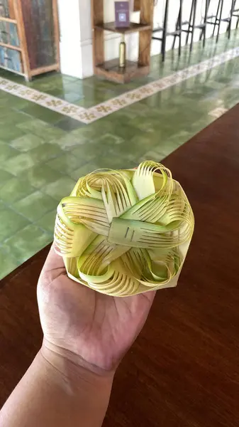 El ele tutuşmak, dokunmuş avuç içi yaprağı süsü. Bir el, palmiye yapraklarından örülmüş kısmen tamamlanmış kare bir süsü tutar. Karmaşık deseni ve işçiliği gösterir..