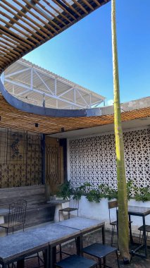 Bambu pergola, geometrik desenli duvar ve metal mobilyalı açık hava kafe..