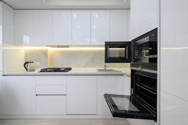 White modern kitchen with a stove, oven and microwave with open doors, glass electric kettle at worktop.
