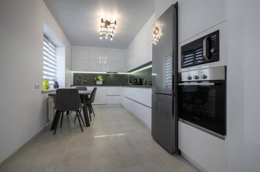 Luxurious modern trendy white and grey kitchen interior after renovation, with granite counter top and marble floor, dining table next to window