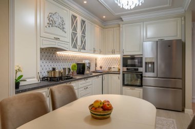 Bej renkli ve klasik tasarım unsurları olan şık, açık fikirli bir mutfak. Meyveli yemek masasına yakın.