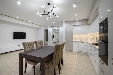 Duvarında televizyon olan büyük, beyaz, lüks bir stüdyo dairenin içinde büyük bir mutfak, bir sürü fotokopi alanı, sandalyeli yemek masasının yakın görüntüsü.