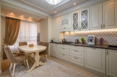 Geniş ve davetkar bej mutfak, yemek masası ve modern aletleri var.