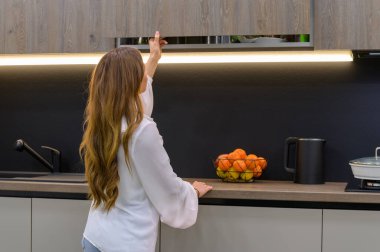 Young and pretty woman opens cabinet door in interior of modern grey large luxury kitchen