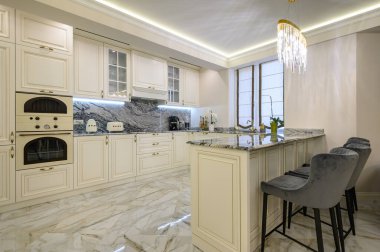 Krem renkli klasik mutfak ve stüdyo dairesinin içinde yemek alanı olarak kullanılan ada, yastıklı ve perdeli rahat kahverengi kanepe