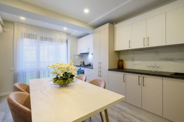 Geniş bir bej ve krema mutfağının içi, orkide çiçekli yemek masasına yakın.