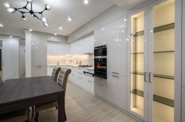 Yemek masası ve sandalyeler, beyaz ferah döşeli mutfak içi fırın ve sağ tarafta büyük LED aydınlanmış büfe.