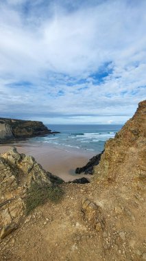 Zambujeira deniz kıyısında deniz dalgaları, uçurumlar ve kumullarla kaplı yeşil bitki örtüsü kırmızı incir yaprakları, güneşli bir gün, açık mavi gökyüzü. Rota Vicentina Sahili, Portekiz.