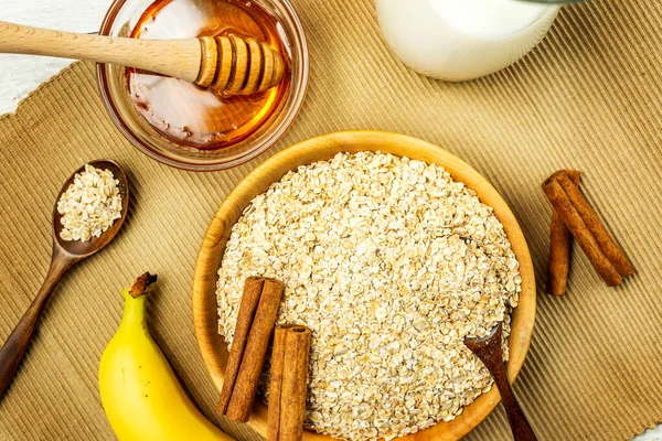 Rolled oats or oat flakes in bowl with wooden spoons, banana, honey, Cinnamon sticks and bottle of milk on background. Healthy lifestyle, healthy eating concept