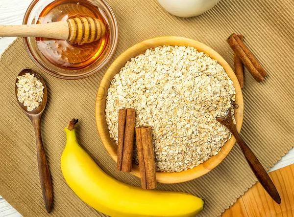 Rolled oats or oat flakes in bowl with wooden spoons, banana, honey, Cinnamon sticks and bottle of milk on background. Healthy lifestyle, healthy eating concept