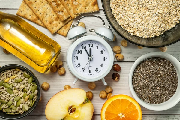Diet time. Food clock. Healthy eating with oat flakes, hazelnuts, chia seeds, pumpkin seeds, sunflower seeds, honey, rye bread a and an alarm clock on a white wooden background