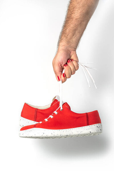 Male hand with red painted fingernails holding hanging red sneakers by the laces on white background. Hand with a new sport shoe.