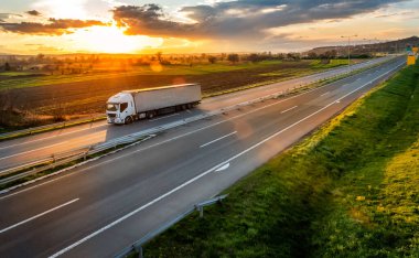 Gün batımında kırsal bir arazide asfalt bir yolda römorklu beyaz nakliye kamyonu.