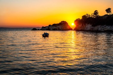 Denizin üzerinde gün batımı. Güneş ufukta iki kayanın arasından batıyor. Yakınlarda küçük bir motorlu tekne var.