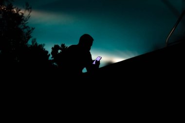Geceleri havuz kenarında oturan ve akıllı telefonuna bakan bir adamın arka silueti. Yumuşak odak, siluet fotoğraf