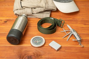 Açık havada ve tahta bir masada yürüyüş malzemesi. Yürüyüş pantolonu, beyzbol şapkası, şişe, pusula, kemer, çakmak ve İsviçre çakısı.