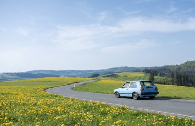blue car under blue sky in spring countryside of german sauerland with trees and blooming fields of dandelions