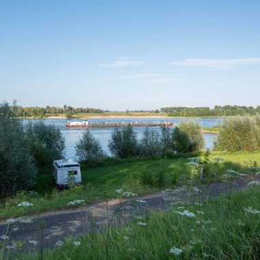 Hollanda 'da Nijmegen yakınlarında nehir kıyısında geçen gemilerle birlikte yaz çiçekleri ve karavan kamyonu.