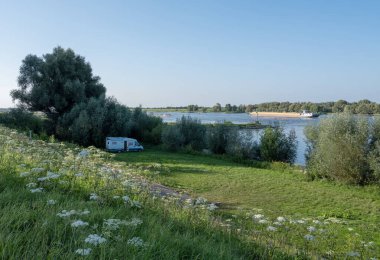 summer flowers and camper van on embankment of river waal near nijmegen in the netherlands with passing ships clipart