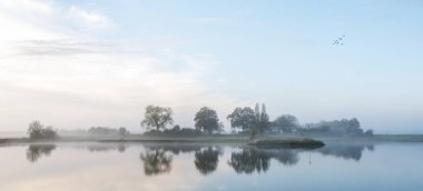 farm and trees reflected in calm water of river lek near wijk bij duurstede in the netherlands on foggy morning in autumn clipart