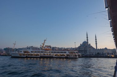İstanbul fotoğrafı. Ferry ve Yeni Cami namı diğer Eminonu bölgesindeki Yeni Cami gün batımında Galata Köprüsü 'nden. İstanbul Türkiye - 12.24.2022