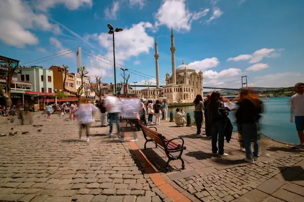 Ortakoy Camii ve turistler. İstanbul konsept fotoğrafını ziyaret edin. Uzun pozlu çekim. İnsanlar ve bulutlar hareket bulanıklığı çekiyor. İstanbul Türkiye - 4.27.2024