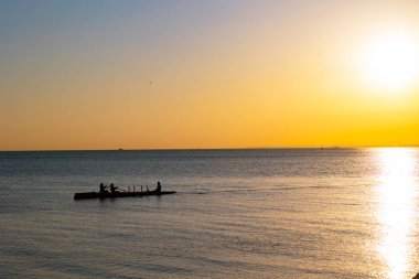 Gün batımında sakin denizin üzerinde kano süren atletlerin siluetleri. Sporcular kanolarını denizde yüzdürüyor..